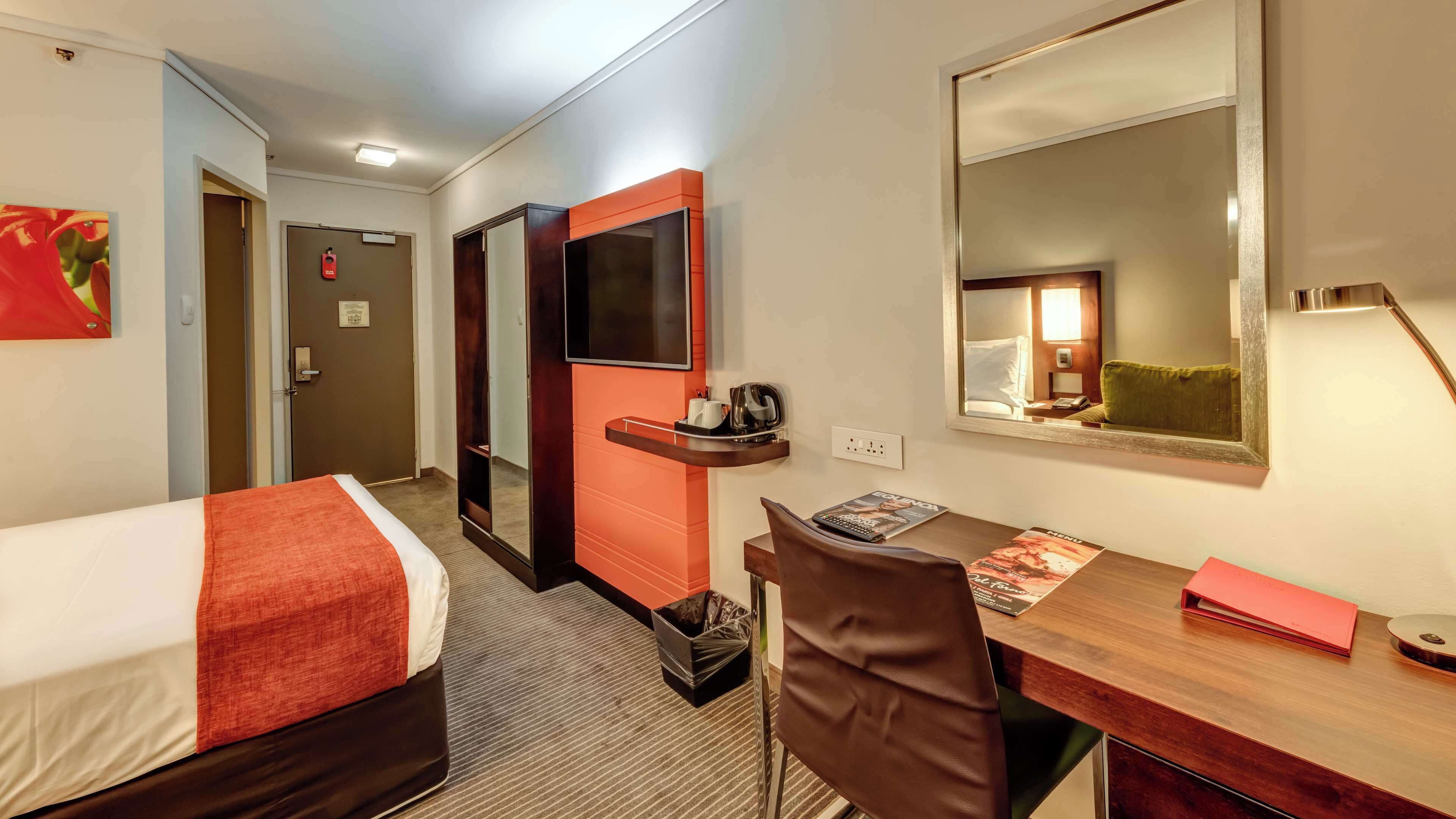 Habitación de hotel con cama, escritorio, silla, espejo, televisor y puerta.