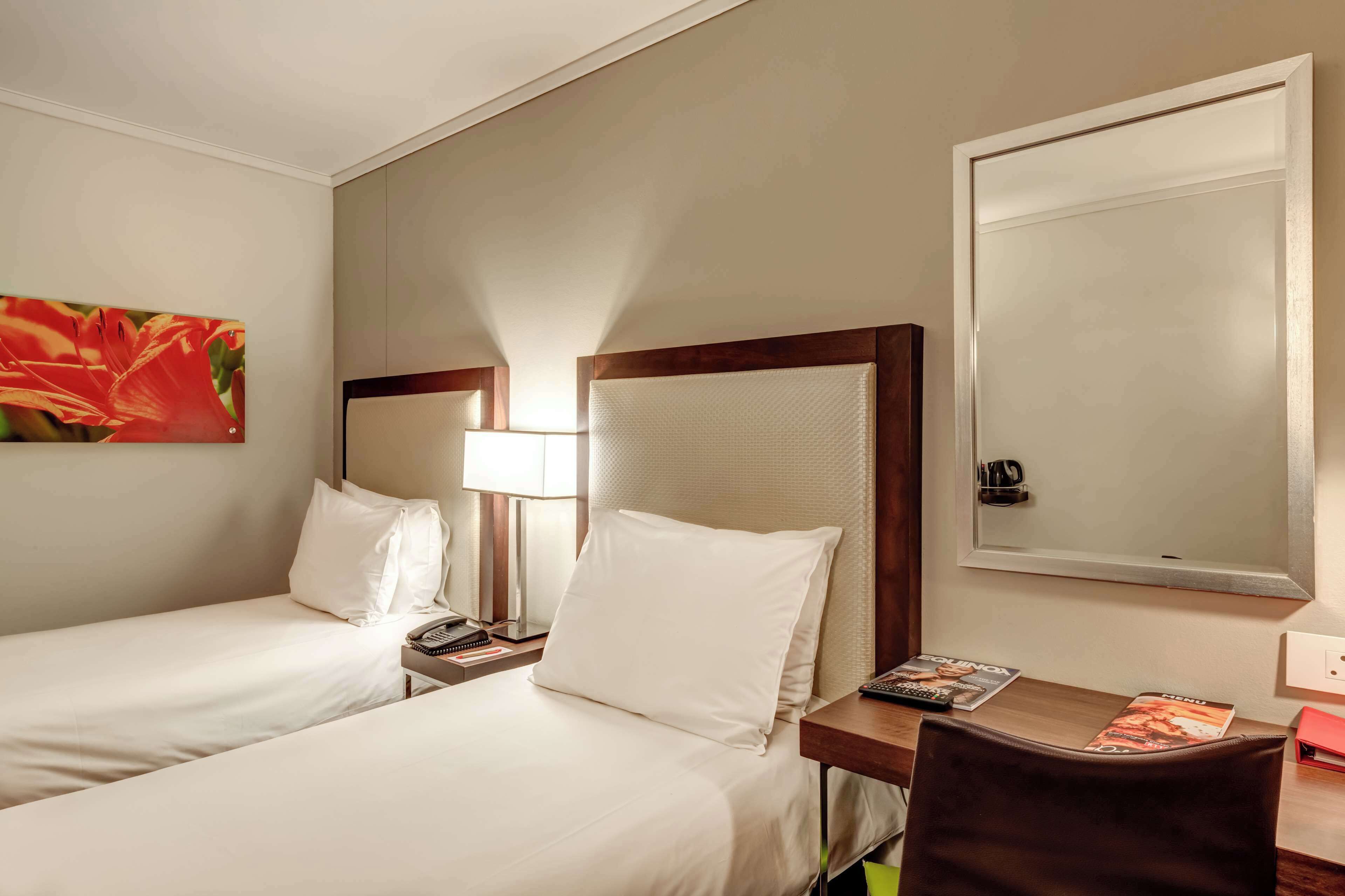 A hotel room with two beds, a desk with a chair, a lamp, a mirror, and a painting on the wall.