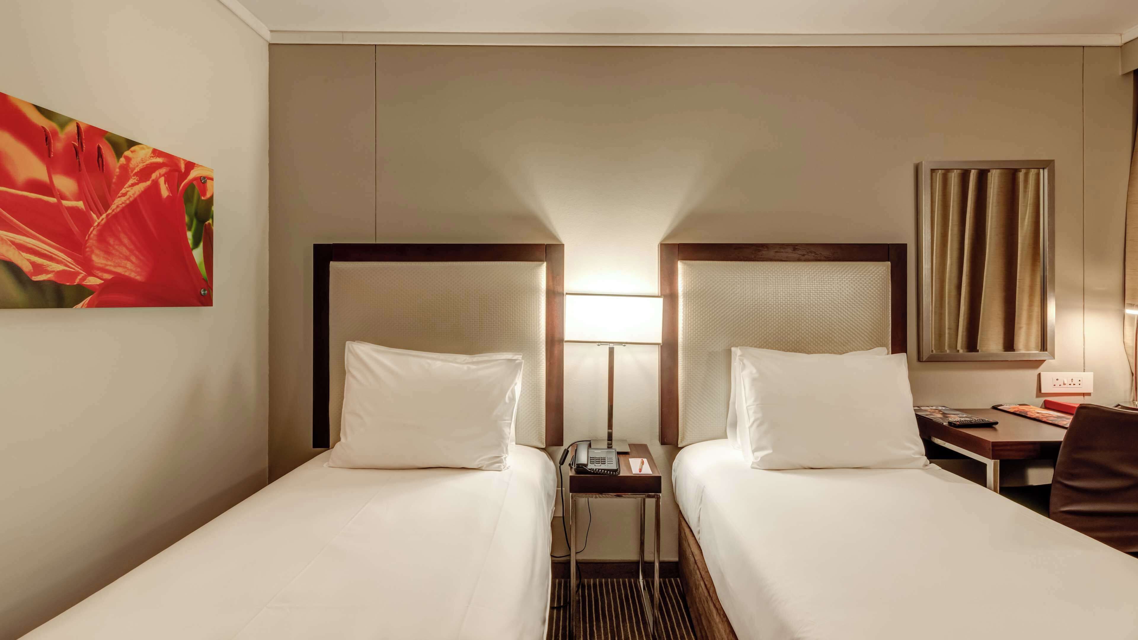 Habitación de hotel con dos camas, una mesita de noche con lámpara, un escritorio con silla y un cuadro en la pared.