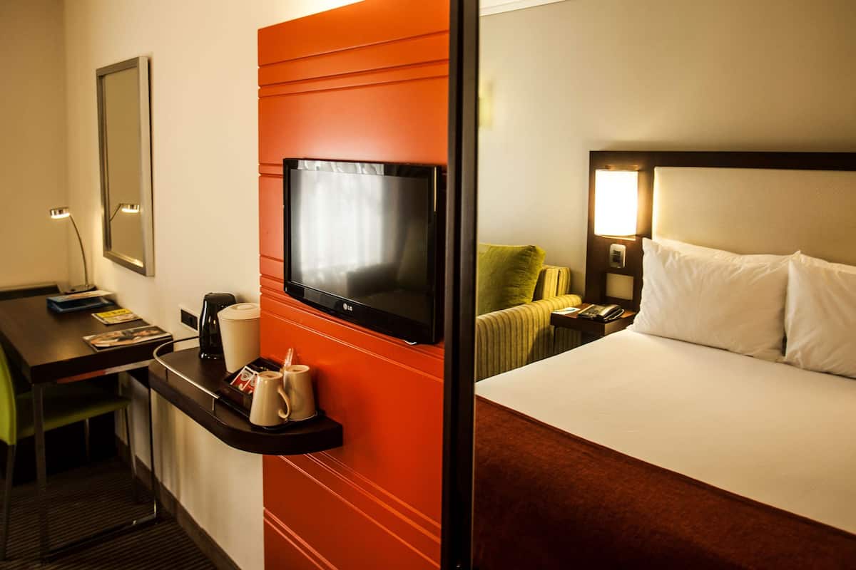 A hotel room with a bed, a desk with a chair, a television, a coffee maker, and a nightstand with a lamp.