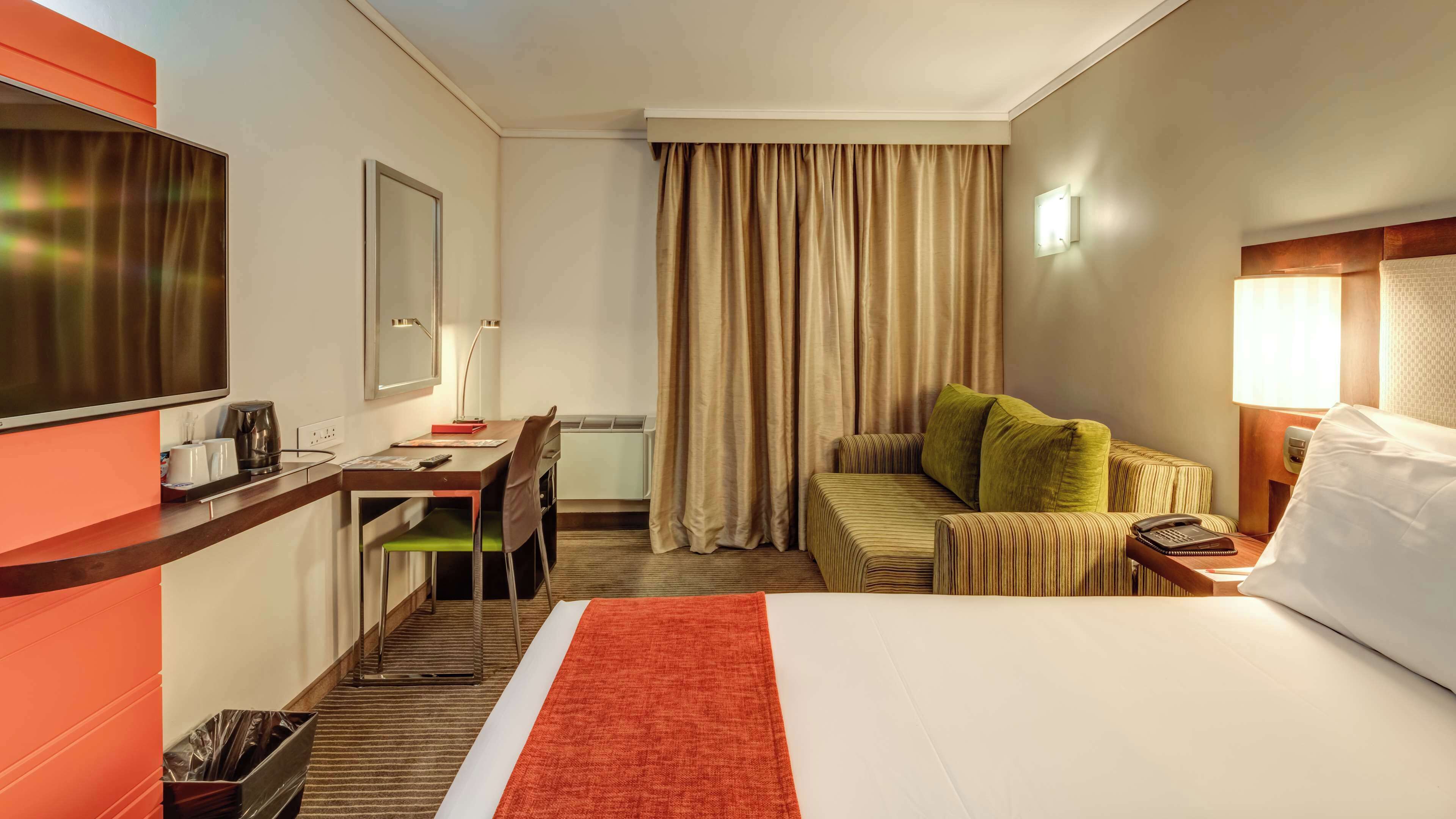 A hotel room with a bed, a desk with a chair, a television, a lamp, and a window with curtains.