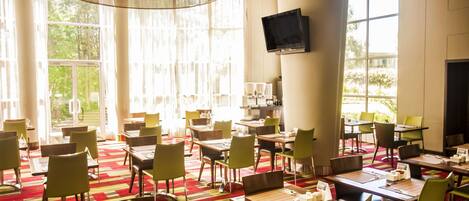 Un comedor de hotel con mesas, sillas, un televisor y ventanas amplias.