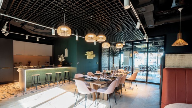 A modern dining area with a long table, multiple chairs, pendant lighting, and a kitchen in the background. - Student Only Brent Cross (London)