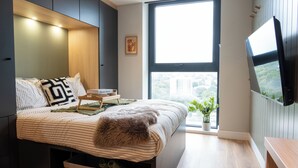 A bedroom with a bed, a TV mounted on the wall, a window with a view, and a small plant. - Student Only Brent Cross (London)