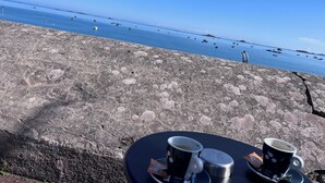 A seaside view with a table set for two, featuring two cups of coffee and a metal container, overlooking a calm sea with boats and a distant island. - RESPIRER A DEUX EN FINISTERE NORD (Taulé)
