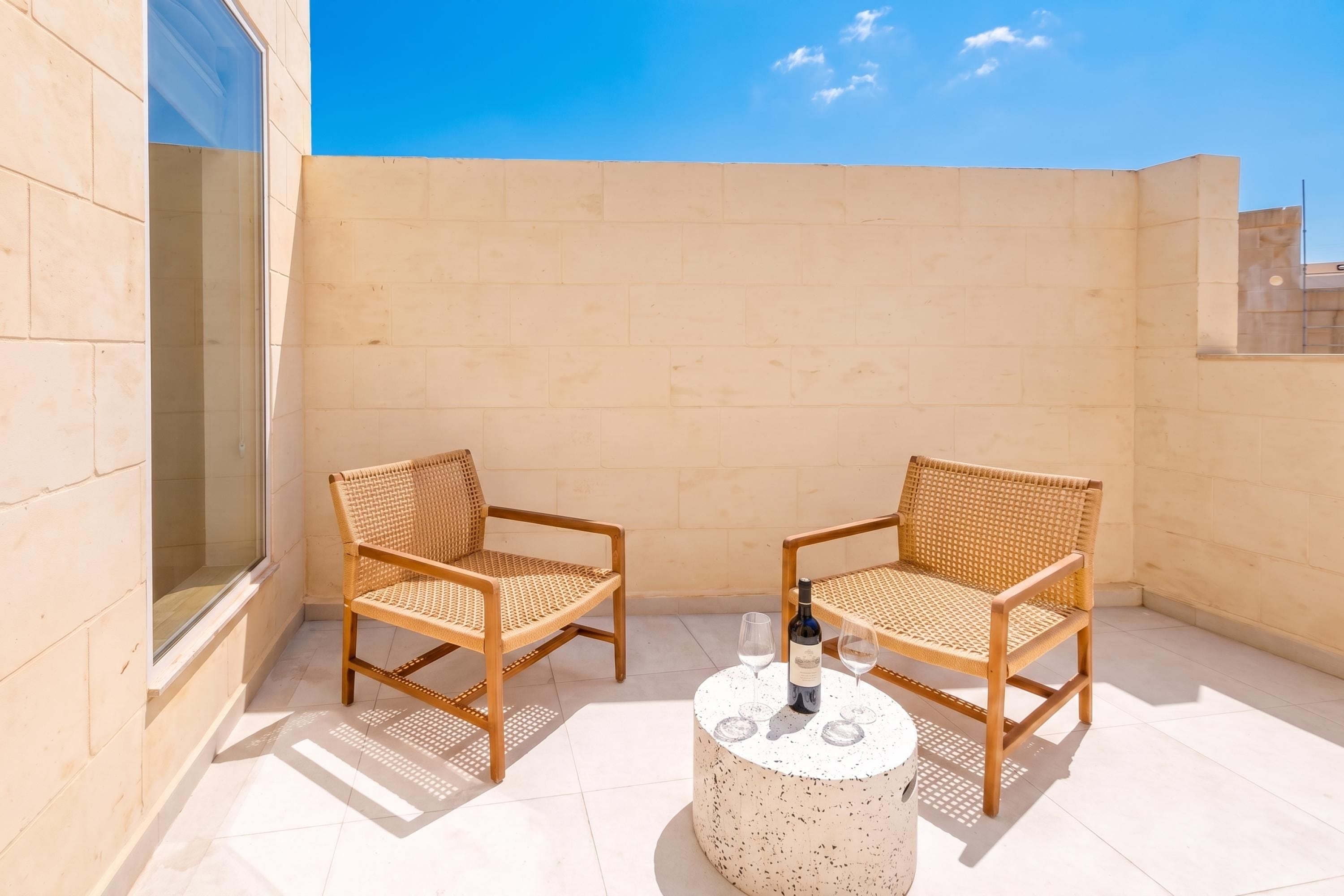 A balcony with two wicker chairs and a round table, a bottle of wine, and two glasses.