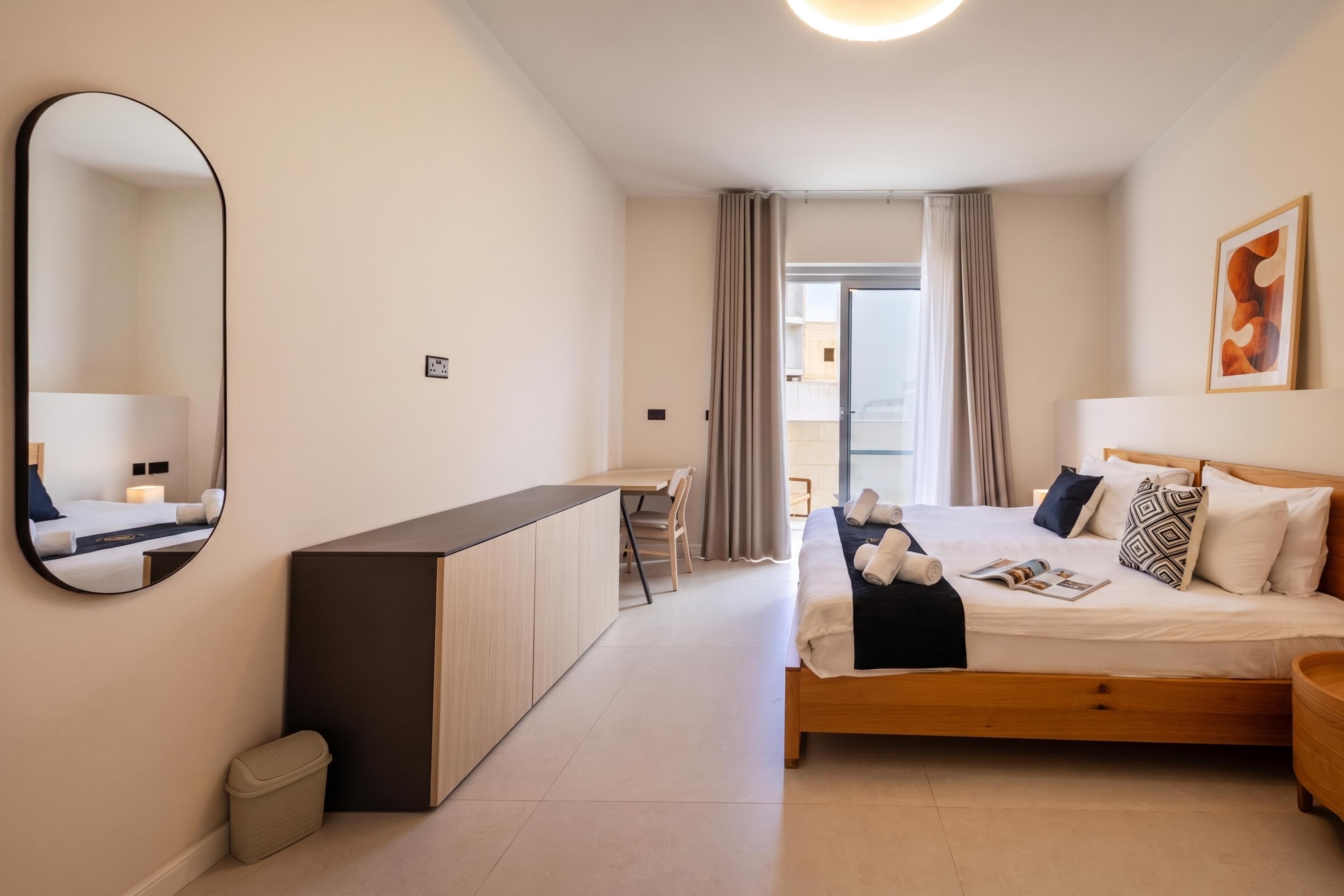 A modern hotel room with a large bed, a bedside table, a mirror, and a view of the outside.