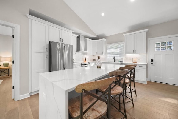 A modern kitchen with white cabinetry, a large island with bar stools, stainless steel appliances, and a window providing natural light.