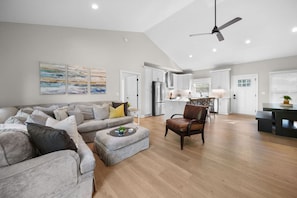 A spacious living room with a grey sofa, a coffee table, and a leather chair. There is a kitchen area with white cabinets and a dining table.