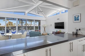 A modern kitchen with a large island, a dining area with a round table and chairs, a living area with a sofa and a TV, and large windows with a view of palm trees. - Beautiful Canyon View Estates Condo with amazing views (Palm Springs)