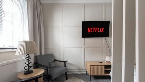 A modern living room with a grey sofa, a small table with a lamp, a wooden side table, and a TV mounted on the wall displaying "NETFLIX".