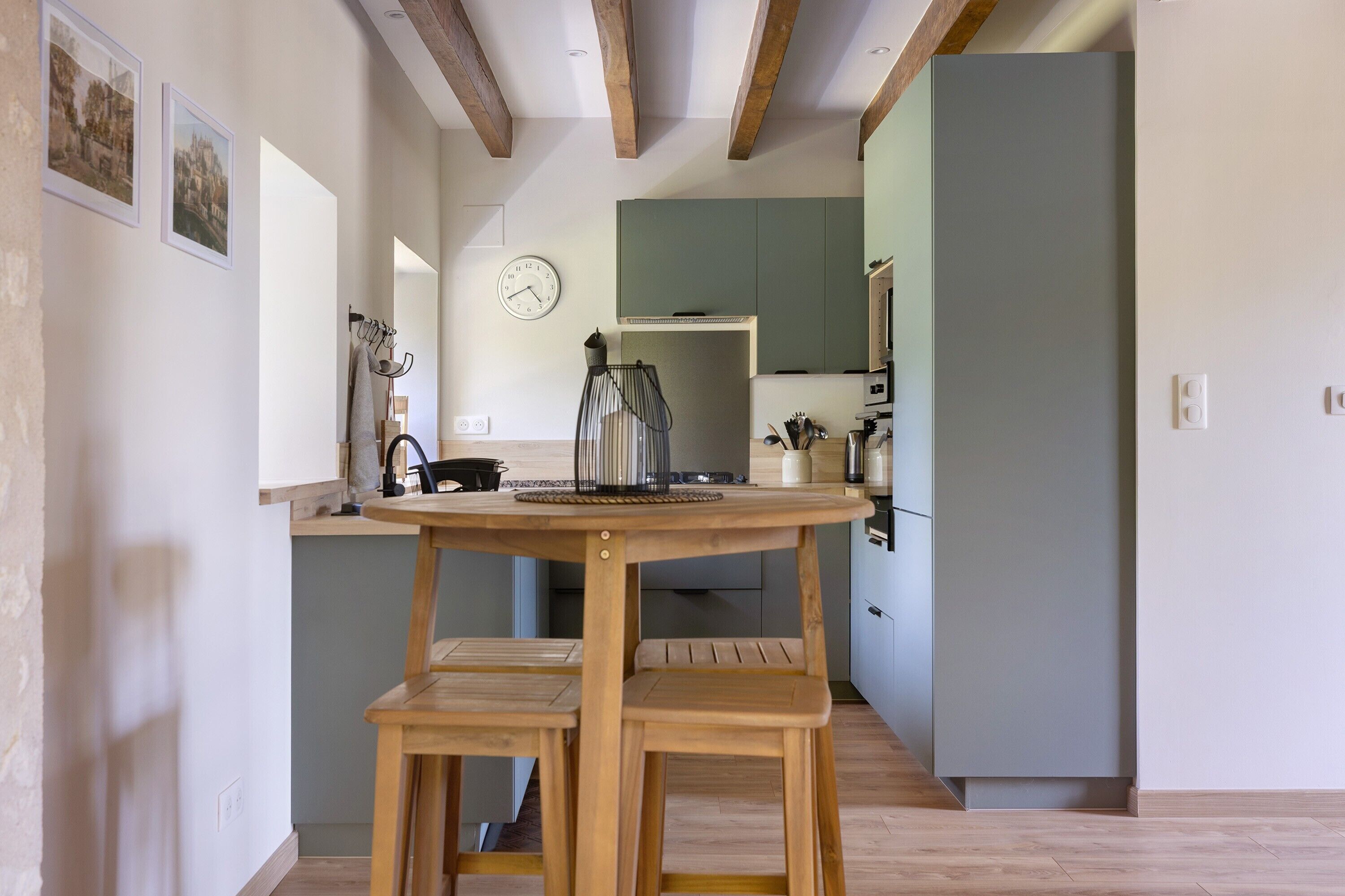 Uma cozinha com mesa e banquetas de madeira, um relógio na parede e armários verdes.