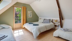 Een slaapkamer met een schuine kap, een raam met gordijnen, een bed met een geruit sprei en een nachtkastje met een lamp.