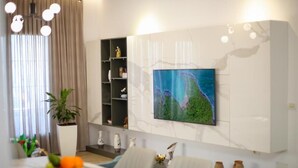 A modern living room with a large TV mounted on the wall, a dining area with a table and chairs, and a potted plant.