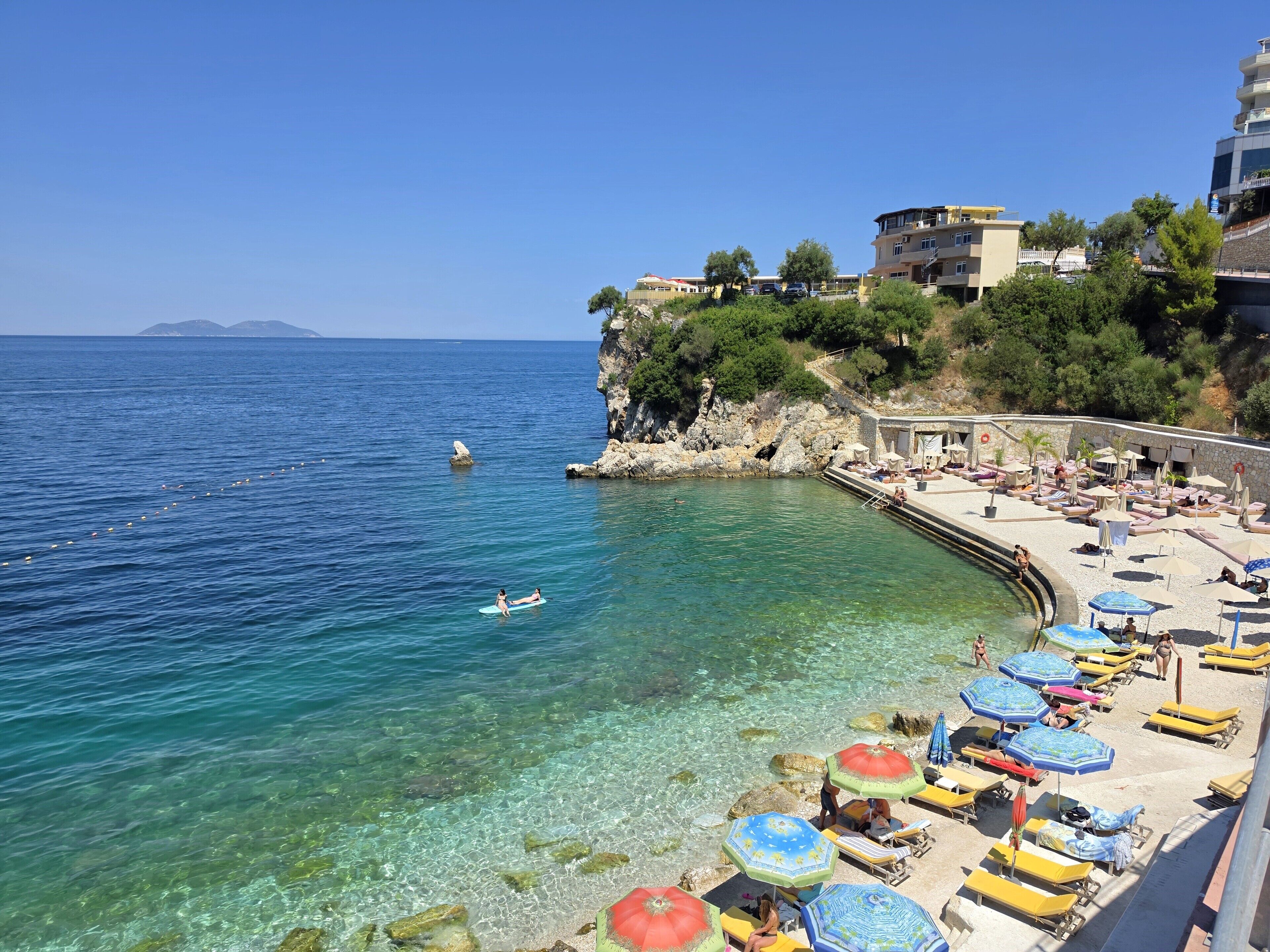 A coastal beach with clear water, sun loungers, umbrellas, and a rocky cliff with buildings.