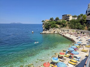 A coastal beach with clear water, sun loungers, umbrellas, and a rocky cliff with buildings.