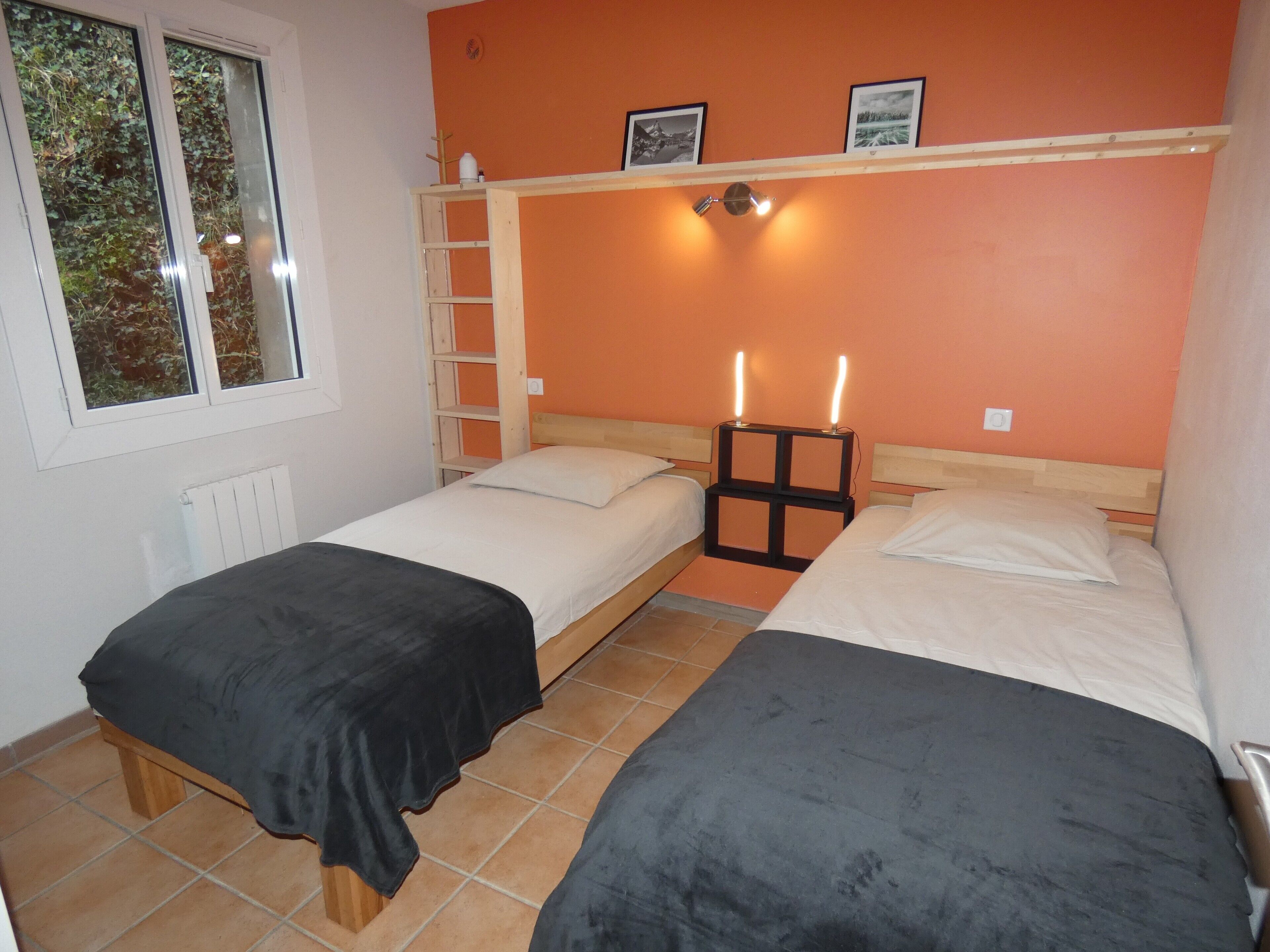 Una habitación con dos camas, una ventana, un radiador y una estantería con fotografías.