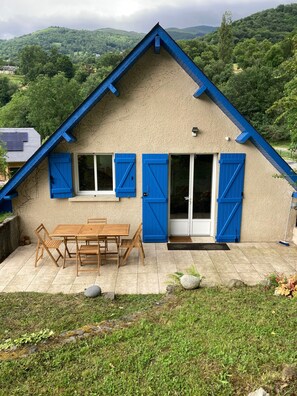 Een huis met een zadeldak, een terras met een houten tafel en stoelen, en uitzicht op een groen landschap.
