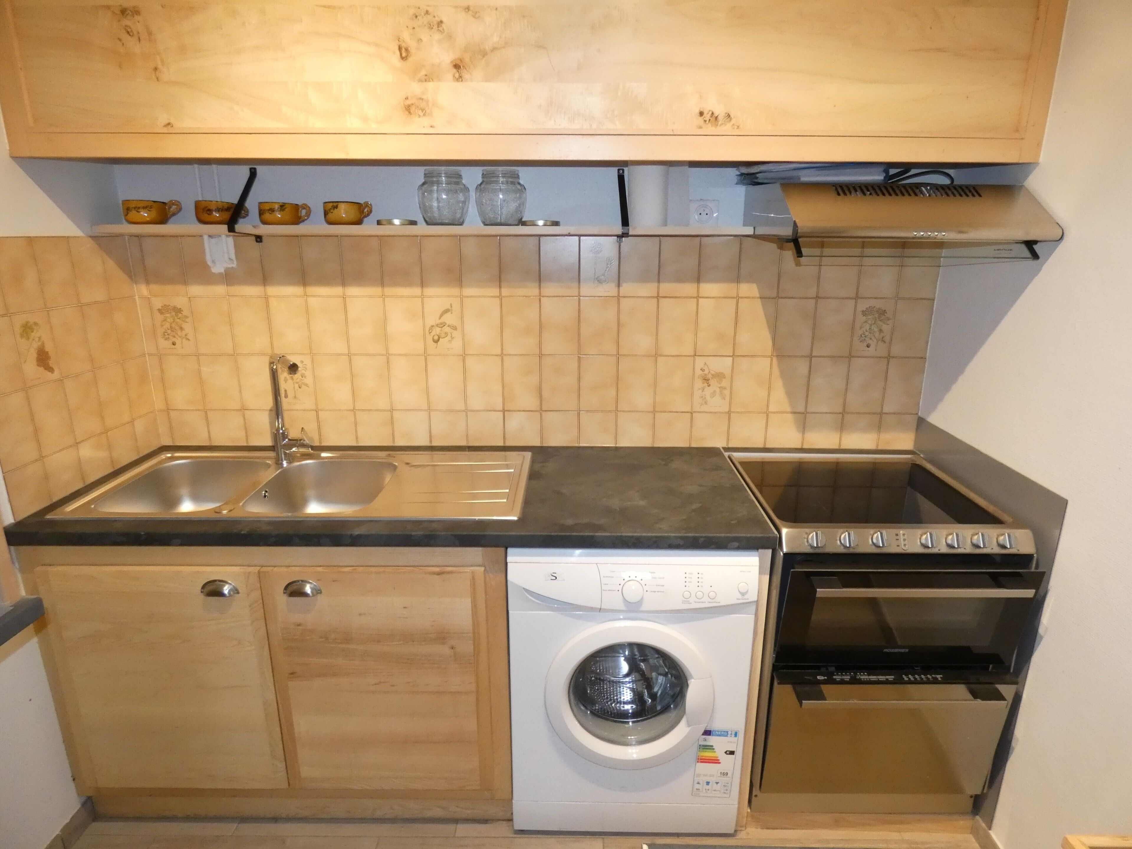 A compact kitchen with a sink, a washing machine, an oven, and wooden cabinets.