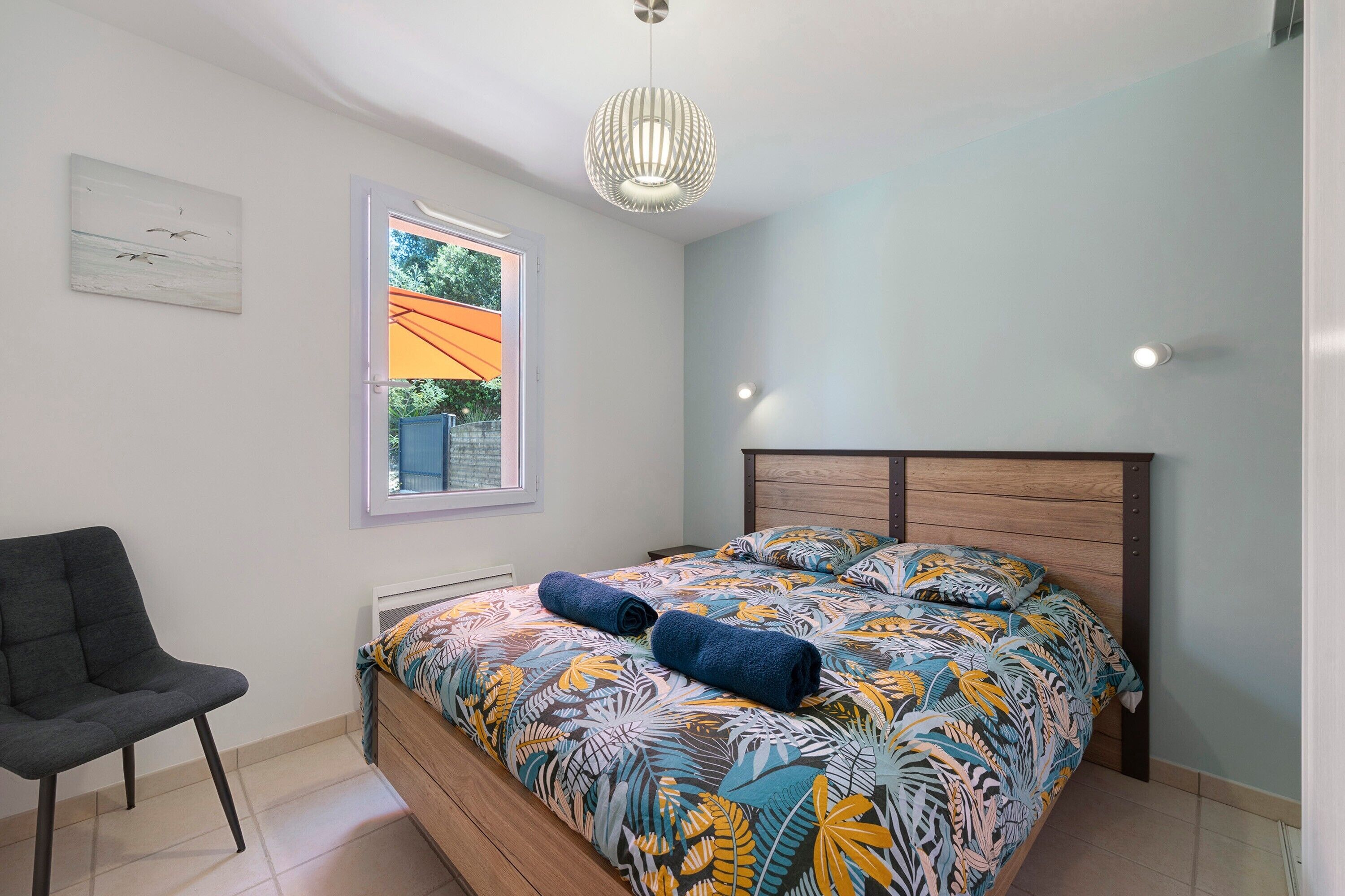 A bedroom with a bed, a chair, a window with an orange awning, and a ceiling light fixture.