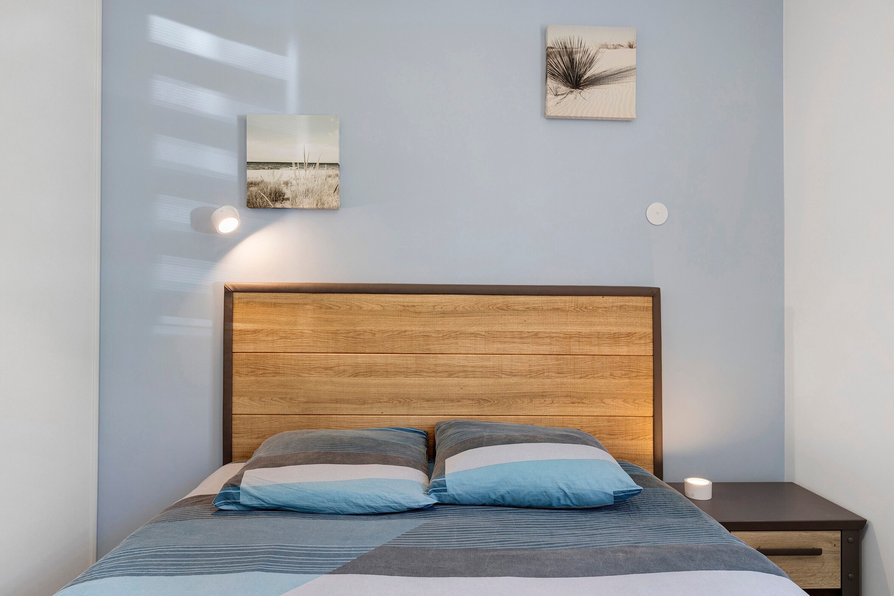 Een bed met houten hoofdbord, twee kussens, een nachtkastje met lamp en twee aan de muur hangende foto's.