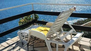 Un balcón con una silla blanca y un cojín amarillo, con vista a un cuerpo de agua y colinas.
