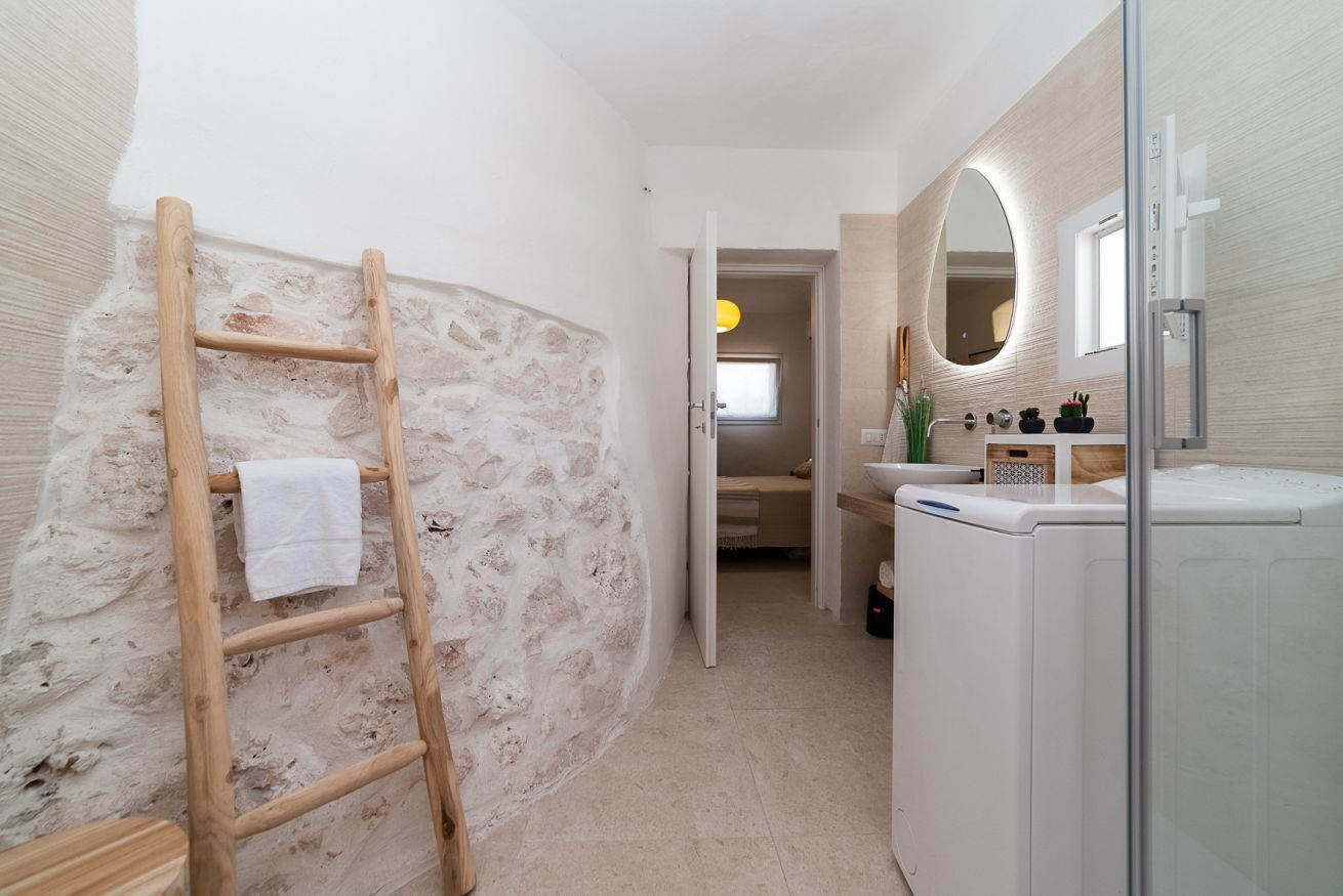 A bathroom with a stone wall, a wooden ladder, a washing machine, a sink with a mirror, and a door leading to another room.