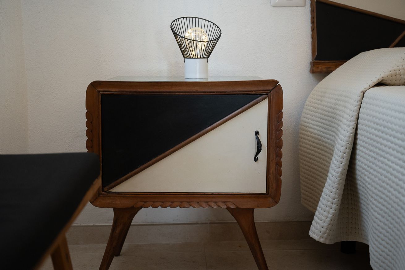 A small bedside table with a lamp, a bed with a white quilt, and a wooden chair.