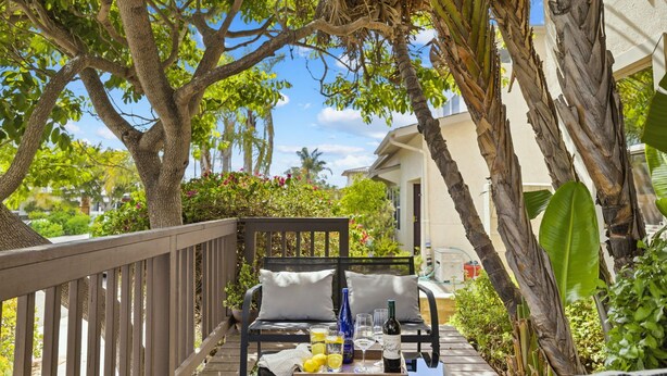 A wooden deck with a black wicker sofa, a tray with drinks and a bottle, surrounded by lush greenery and a house in the background. - Mission Bay Canyon House | Spanish View Retreat (San Diego)