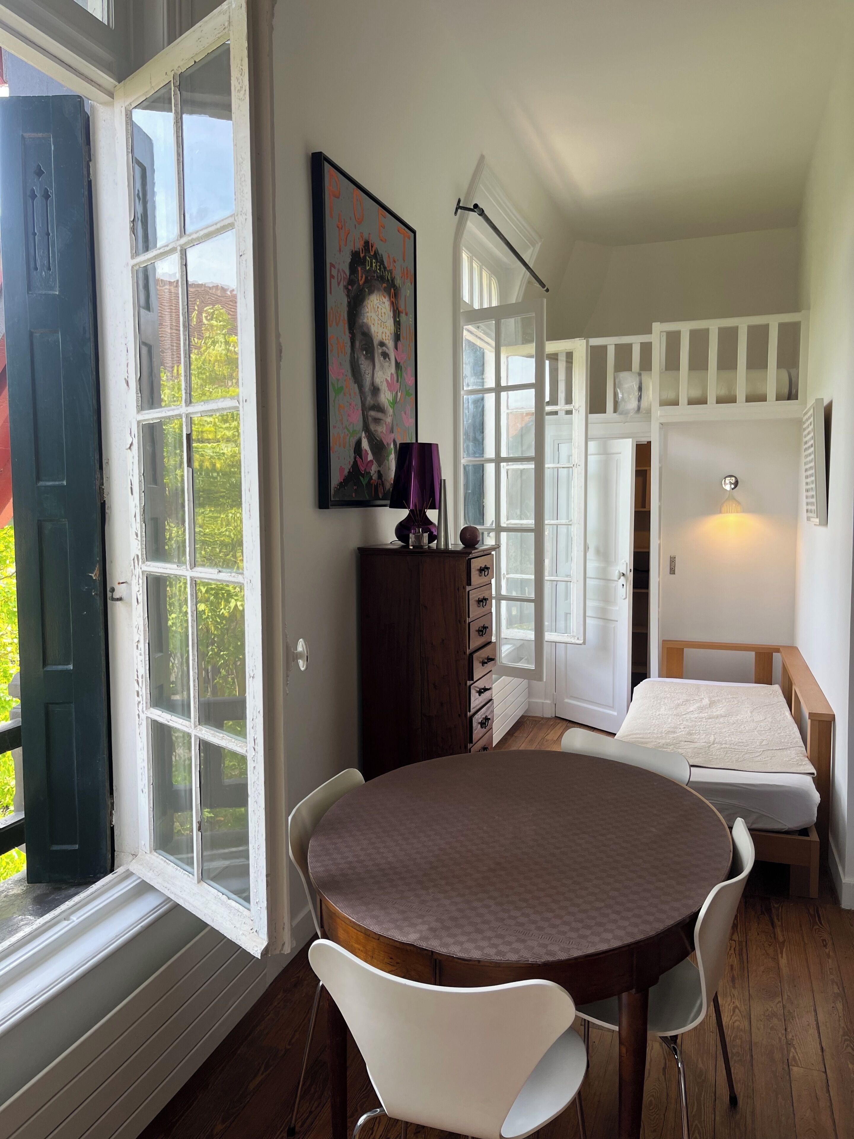 A room with a round table, white chairs, a bed, a dresser, and a painting.