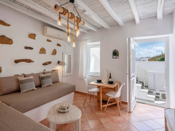 Ein modernes Wohnzimmer mit einer Couch, einem Esstisch und Stühlen, einem kleinen runden Tisch mit einem Glas sowie Blick durch eine offene Tür.