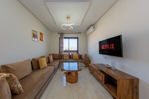 A modern living room with a glass-top coffee table, a sofa set, a wooden sideboard, and a mounted TV displaying the Netflix logo. - Apartment in the center of Guéliz with swimming pool (Marrakesh)