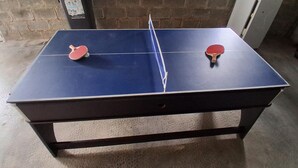Un tavolo da ping pong con due racchette, un pavimento strutturato e un muro in mattoni.