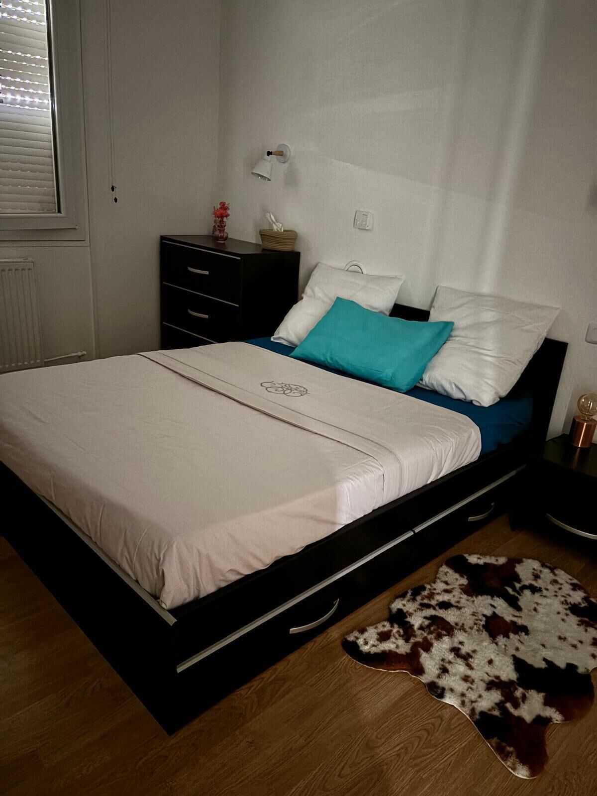Een slaapkamer met een bed, nachtkastje, lamp, dressoir en een vloerkleed van leer.