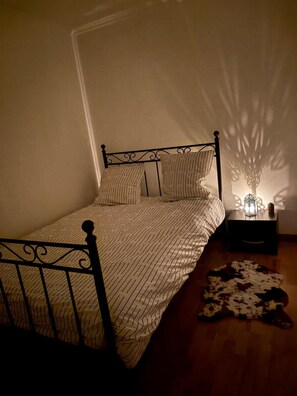 A bed with a metal frame, a patterned bedspread, and pillows. There is a small bedside table with a lamp and a decorative item. A cowhide rug is on the floor. - At Tati Mag (Thiberville)