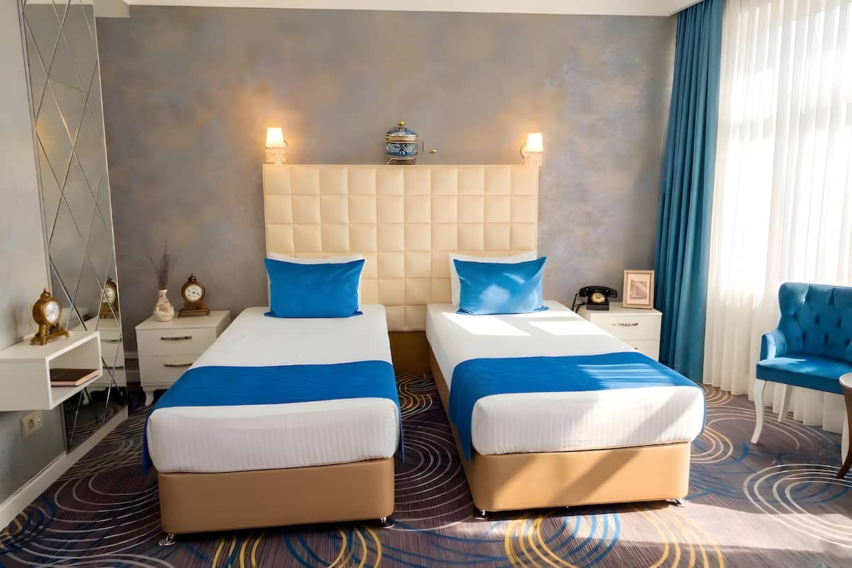 Une chambre d’hôtel avec deux lits, une grande tête de lit rembourrée, des tables de chevet avec des lampes, une chaise et un élément décoratif mural.