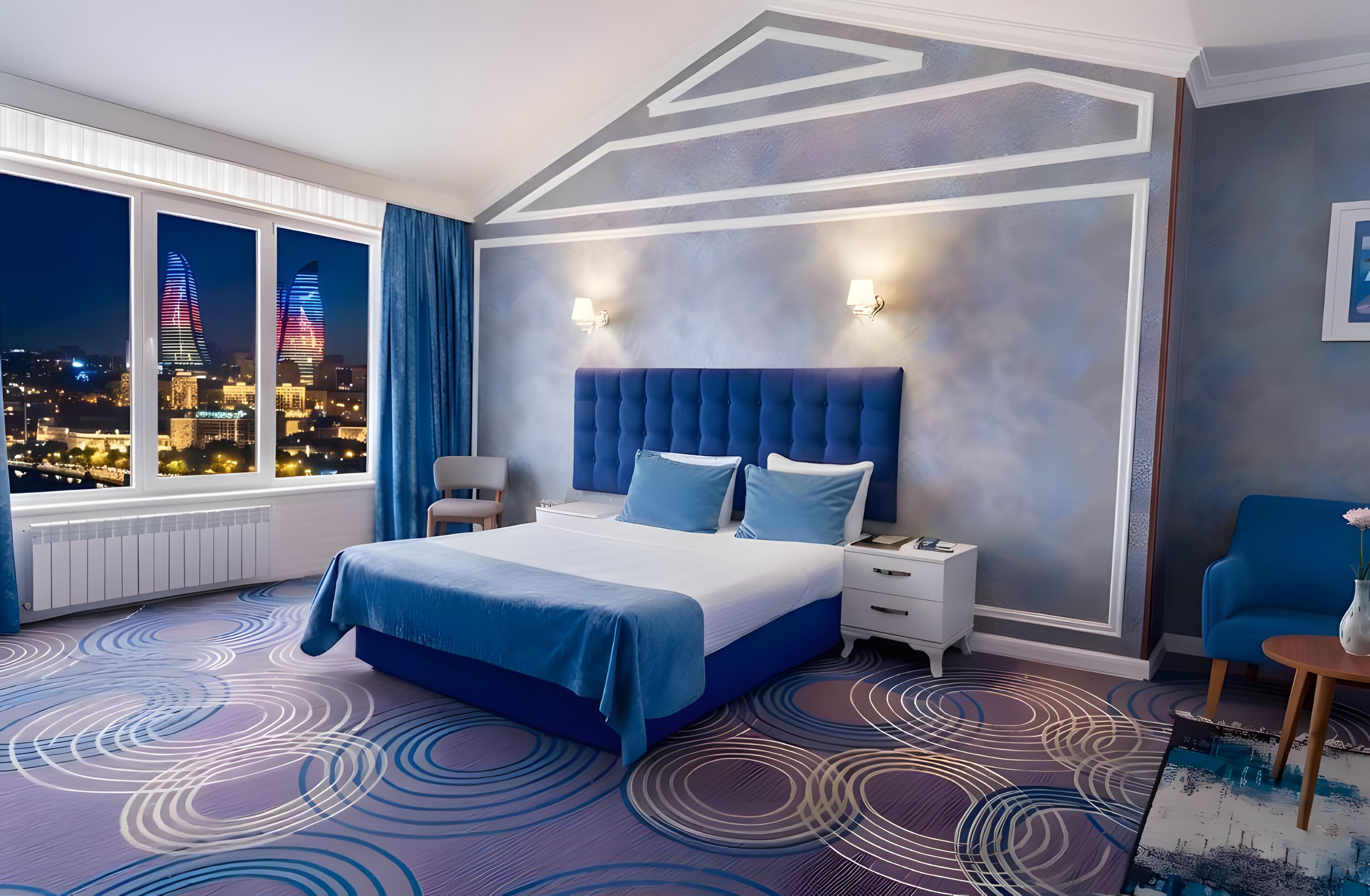 Ein modernes Hotelzimmer mit einem großen Bett, einem Nachttisch, einem Sessel und Blick auf Stadtgebäude sowie einem gemusterten Teppich.