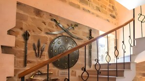 A staircase with a wooden handrail and black wrought iron balusters, leading to a landing with a stone wall and mounted medieval weapons. - Gold Senator Hotel (Baku)