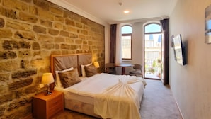 A hotel room with a stone wall, a bed with a headboard, a bedside table with a lamp, and a view of the outside through a window. - Gold Senator Hotel (Baku)