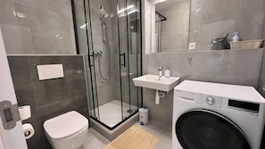 Un baño moderno con ducha separada por mampara de vidrio, inodoro, lavamanos y lavadora.