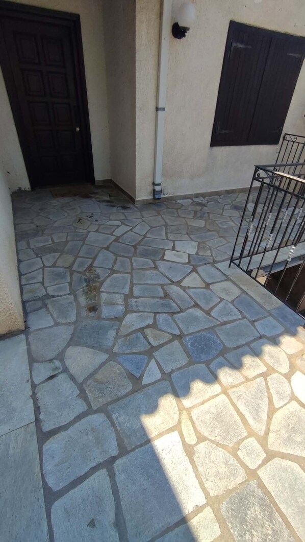 A stone-paved courtyard with a black door, a white pipe, and a metal railing.