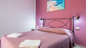 A bedroom with a pink wall, a bed with a metal headboard, two bedside lamps, and a painting of a beach. - Room 'Perla Marina - Scala Dei Turchi' with Balcony, Wi-Fi, Air Conditioning and Shared Kitchen (Realmonte)