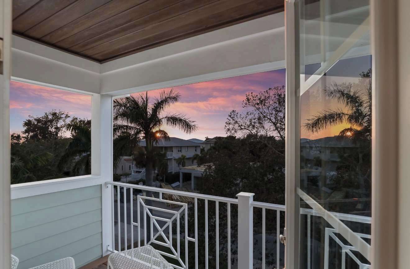 A covered balcony with a view of a sunset, palm trees, and houses.