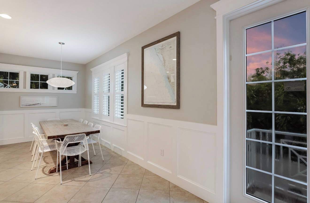 A dining area with a wooden table and white chairs, a large window with blinds, and a framed map on the wall.
