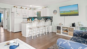 A modern kitchen with white cabinets, a central island, and a dining area with white chairs. - 1BR Condo w/ Pools, Balcony Views & Easy Beach Access | Pebble Beach (Emerald Isle)