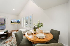 Un coin repas avec une table ronde en bois, des chaises à revêtement vert, une plante en pot, une bouteille de vin et un téléviseur fixé au mur.