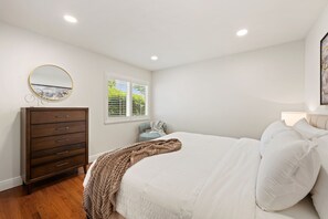 Une chambre à coucher comprenant un lit, une commode, un miroir rond, une fenêtre avec des stores et une lampe de chevet.