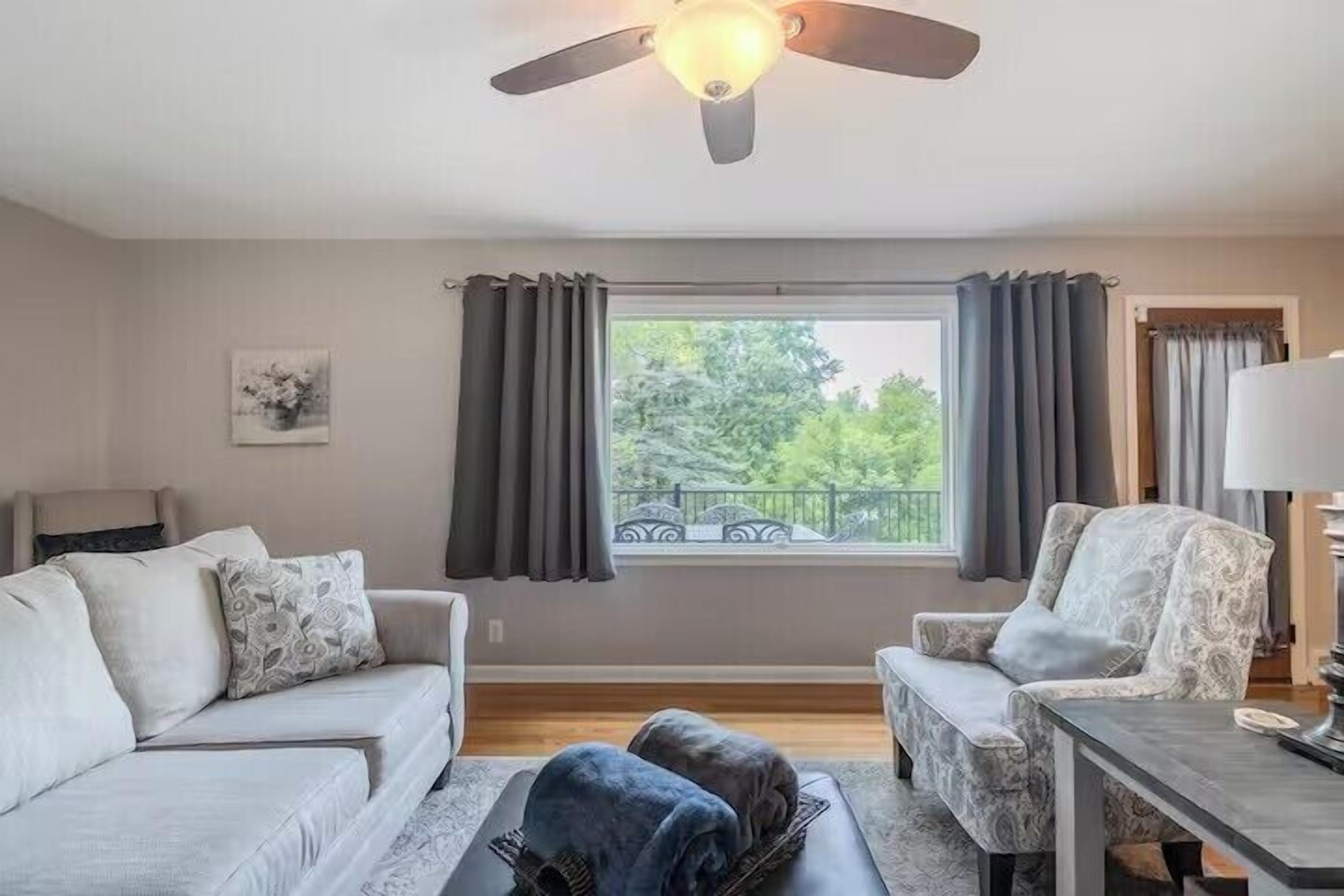 A living room with two sofas, a coffee table, a ceiling fan, a window with curtains, and a view of greenery outside.