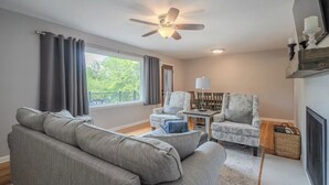 Ein Wohnzimmer mit grauem Sofa, gemusterten Sesseln, einem Deckenventilator, einem kleinen Tisch mit Lampe und einem an der Wand befestigten Fernseher.