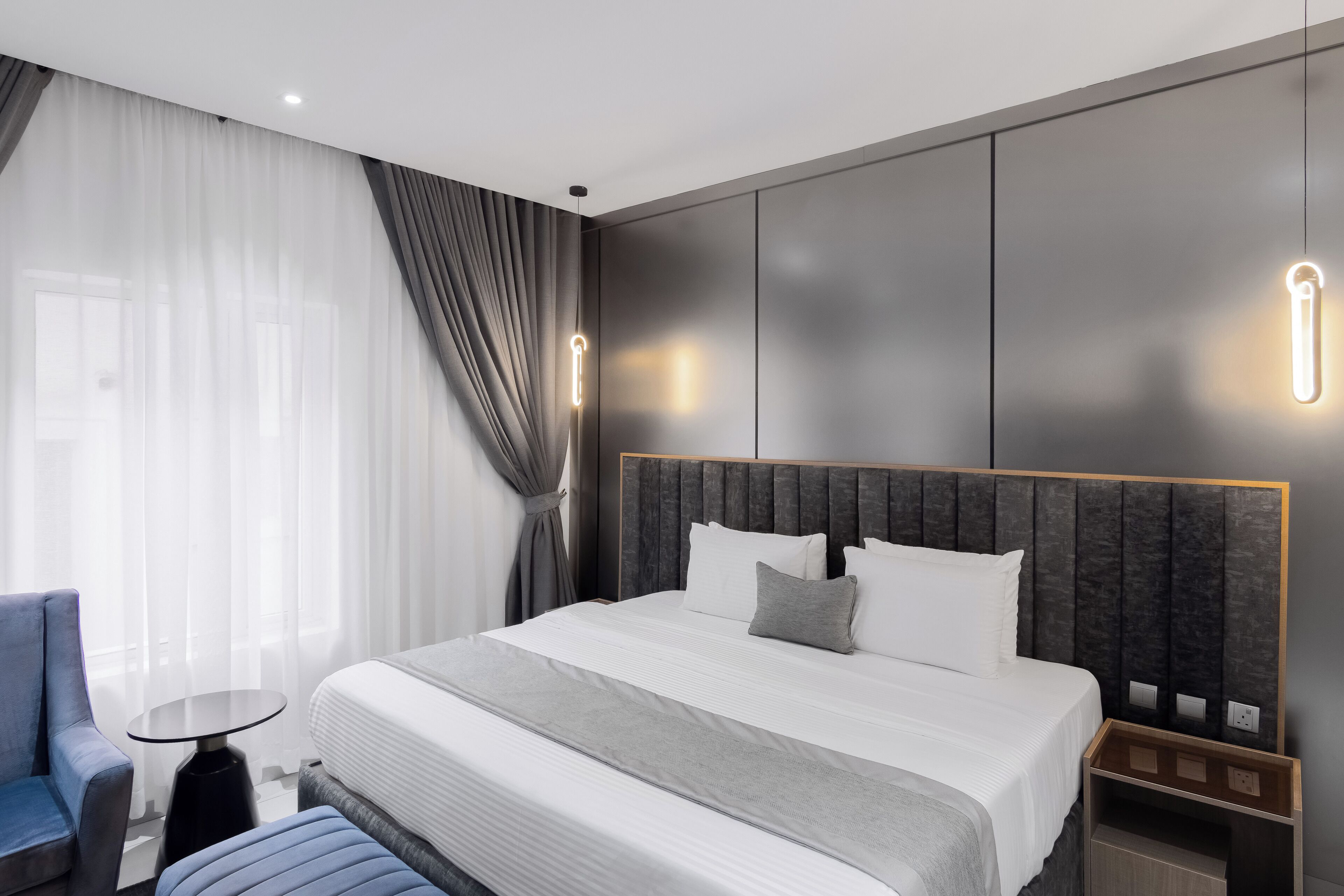 A modern hotel room with a large bed, a small round table, a blue chair, and a nightstand with a lamp.
